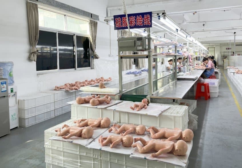 The atelier's production floor: rows of unfinished doll bodies laid out on white crates beside a long assembly line.