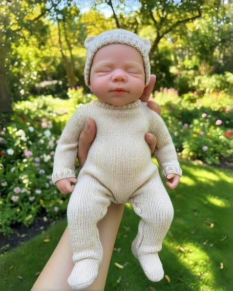 Sleeping reborn doll in a cream knit suit and bear-ear hat, held up by a hand in a sunny garden.