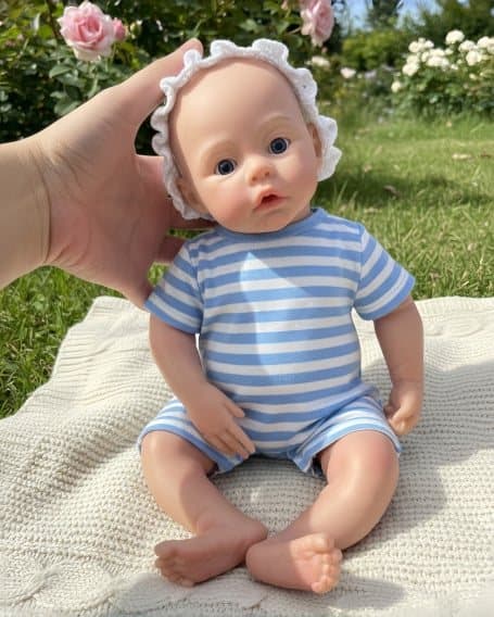 Reborn doll in a blue-and-white striped romper and ruffled bonnet, held upright by a hand in a rose garden.
